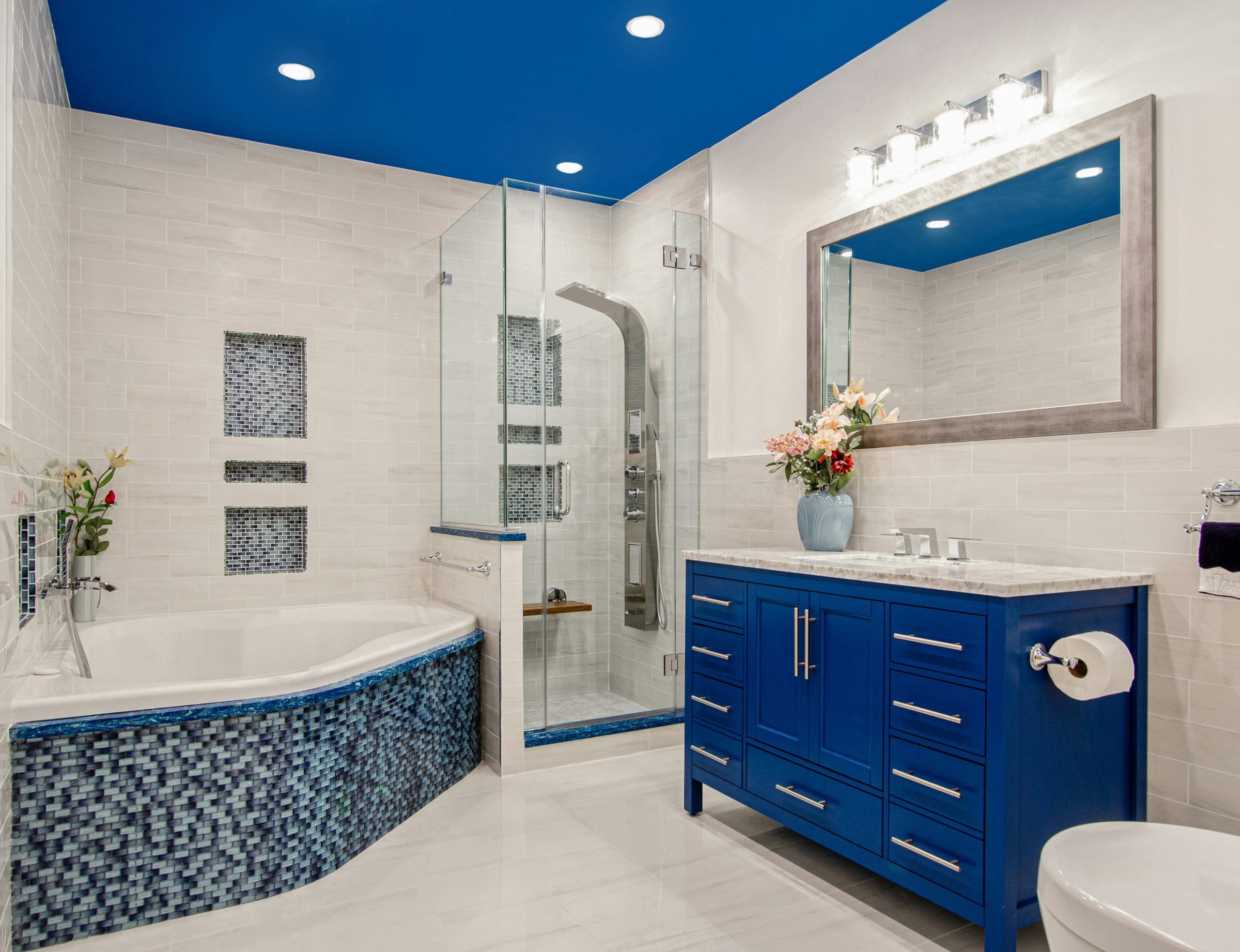 Modern bathroom with a blue ceiling, glass shower, bathtub with blue mosaic tiles, blue vanity with marble countertop, large mirror, and bright lighting; flowers and toiletries decorate the space—expertly finished by a skilled Painter and Decorator.