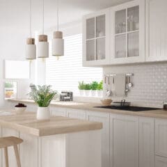 Modern kitchen with light wood cabinets, island with stools, pendant lights, a built-in fridge, sink, stovetop, and various kitchen items on the countertops. Bright natural light streams through a large sash window, recently restored with expert sash window repairs.