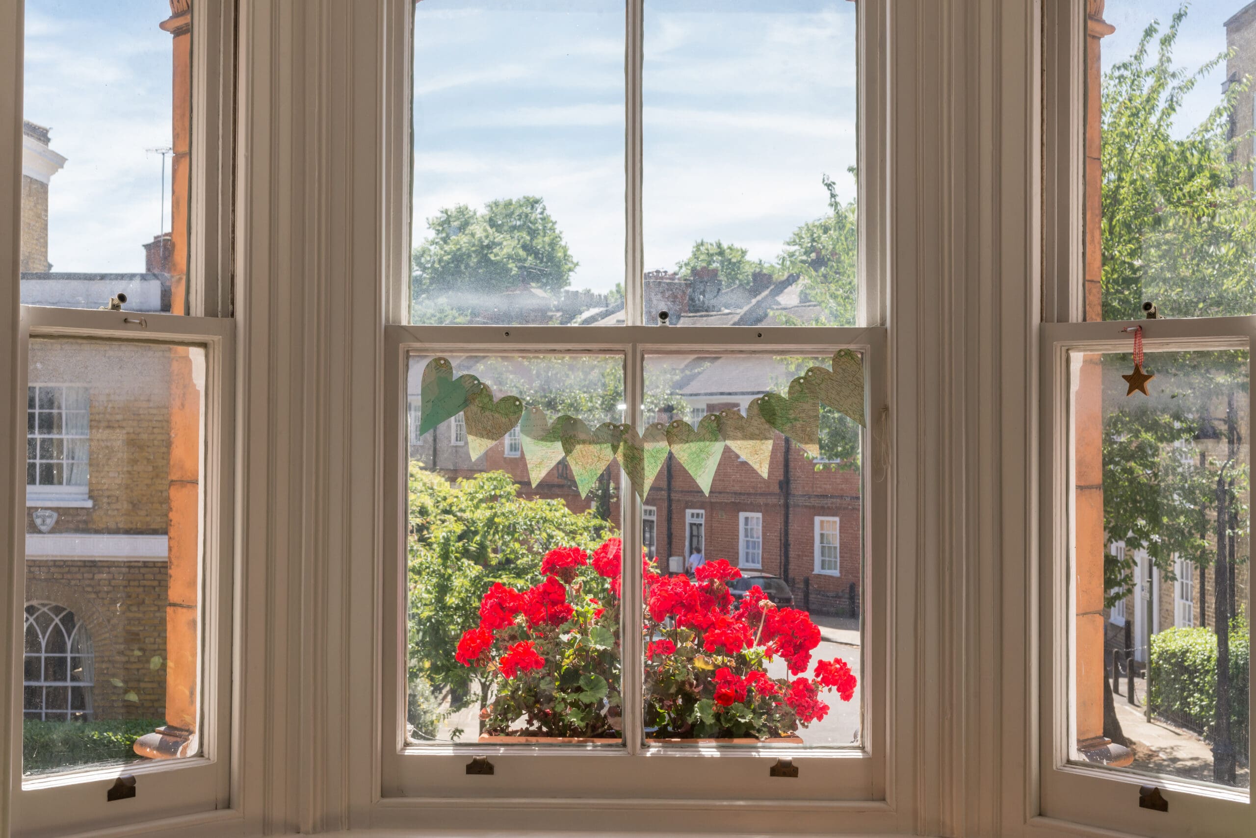 A bay window with a view of a sunny street, red flowers in a planter on the windowsill, green heart-shaped bunting inside, and freshly restored sash window repairs framing trees and brick buildings outside.