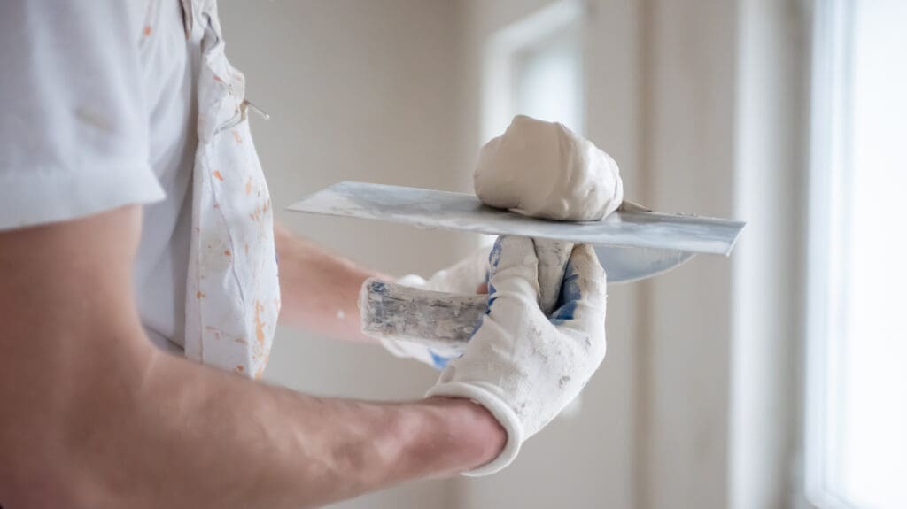 A person wearing gloves and a paint-splattered shirt holds a putty knife with plaster, preparing to apply it to a wall as part of property maintenance in a bright, unfinished room.