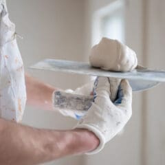 A person wearing gloves and a paint-splattered shirt holds a putty knife with plaster, preparing to apply it to a wall as part of property maintenance in a bright, unfinished room.