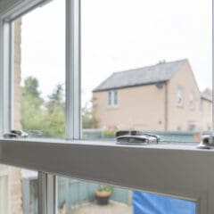 A close-up view of a white, double-glazed sash window, partially open with metal locks visible. Outside, a brick house and greenery can be seen—ideal for those interested in sash window repairs or general property maintenance on an overcast day.