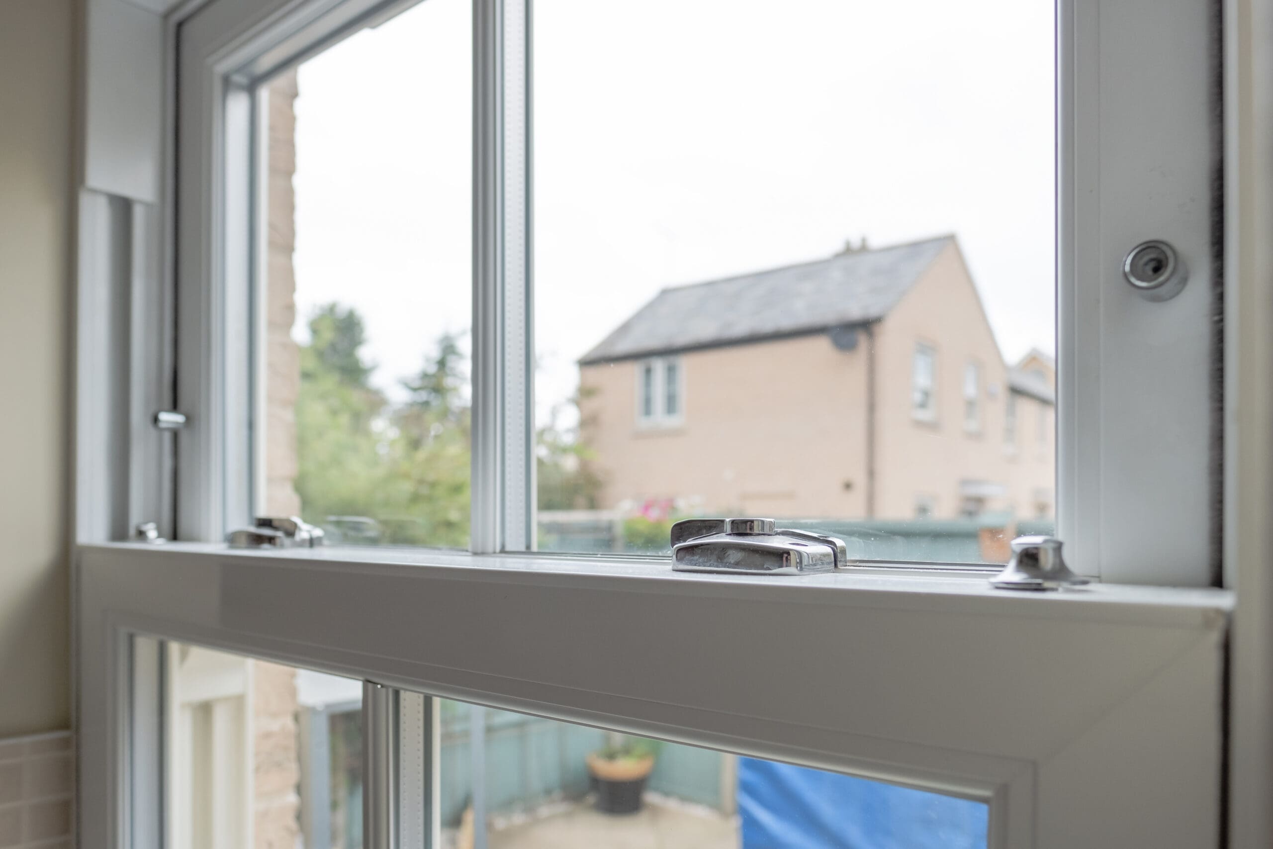 A close-up view of a white, double-glazed sash window, partially open with metal locks visible. Outside, a brick house and greenery can be seen—ideal for those interested in sash window repairs or general property maintenance on an overcast day.