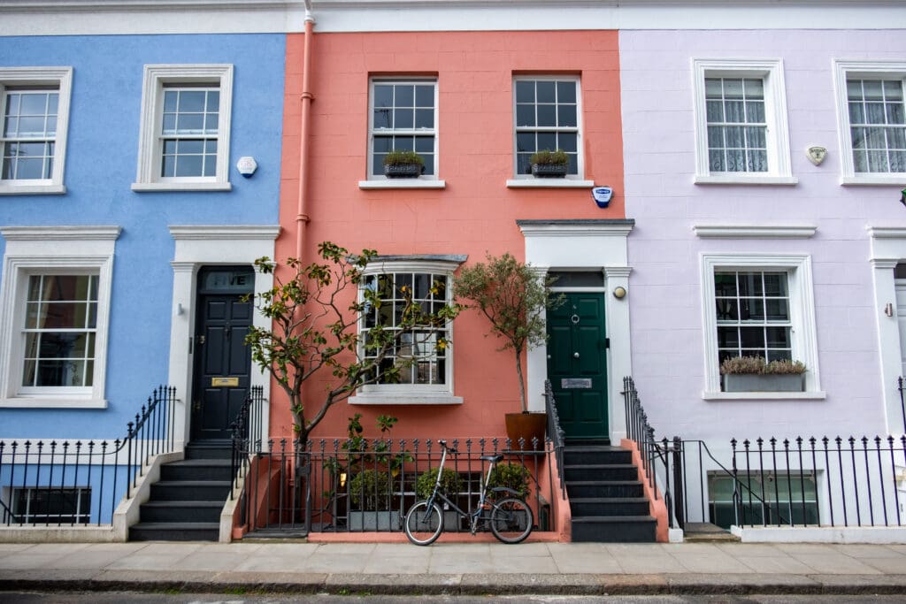 Colorful row houses with blue, coral, and light pink facades, each with black doors and steps. A bicycle is parked in front of the coral house, which features potted plants—a testament to regular property maintenance and care.
