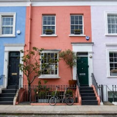 Colorful row houses with blue, coral, and light pink facades, each with black doors and steps. A bicycle is parked in front of the coral house, which features potted plants—a testament to regular property maintenance and care.