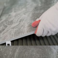 A person wearing white gloves places a large gray tile onto a surface with adhesive, using a plastic spacer to ensure even spacing—demonstrating the precision of a skilled Painter and Decorator at work.