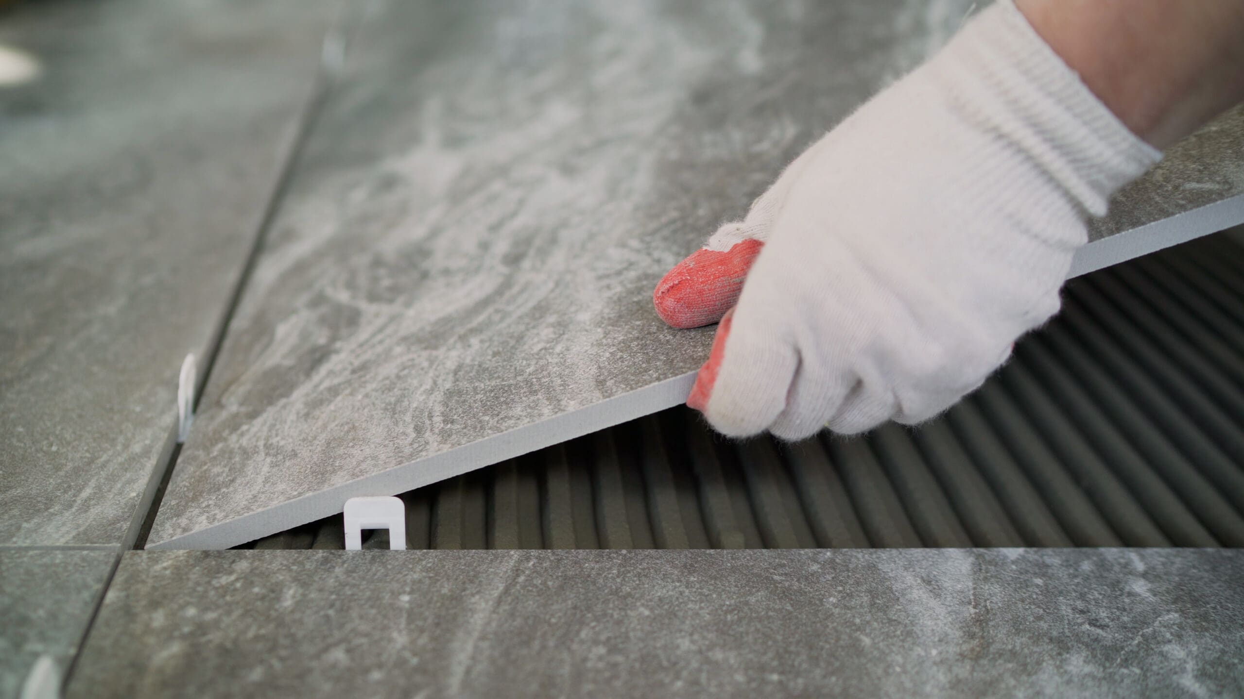 A person wearing white gloves places a large gray tile onto a surface with adhesive, using a plastic spacer to ensure even spacing—demonstrating the precision of a skilled Painter and Decorator at work.