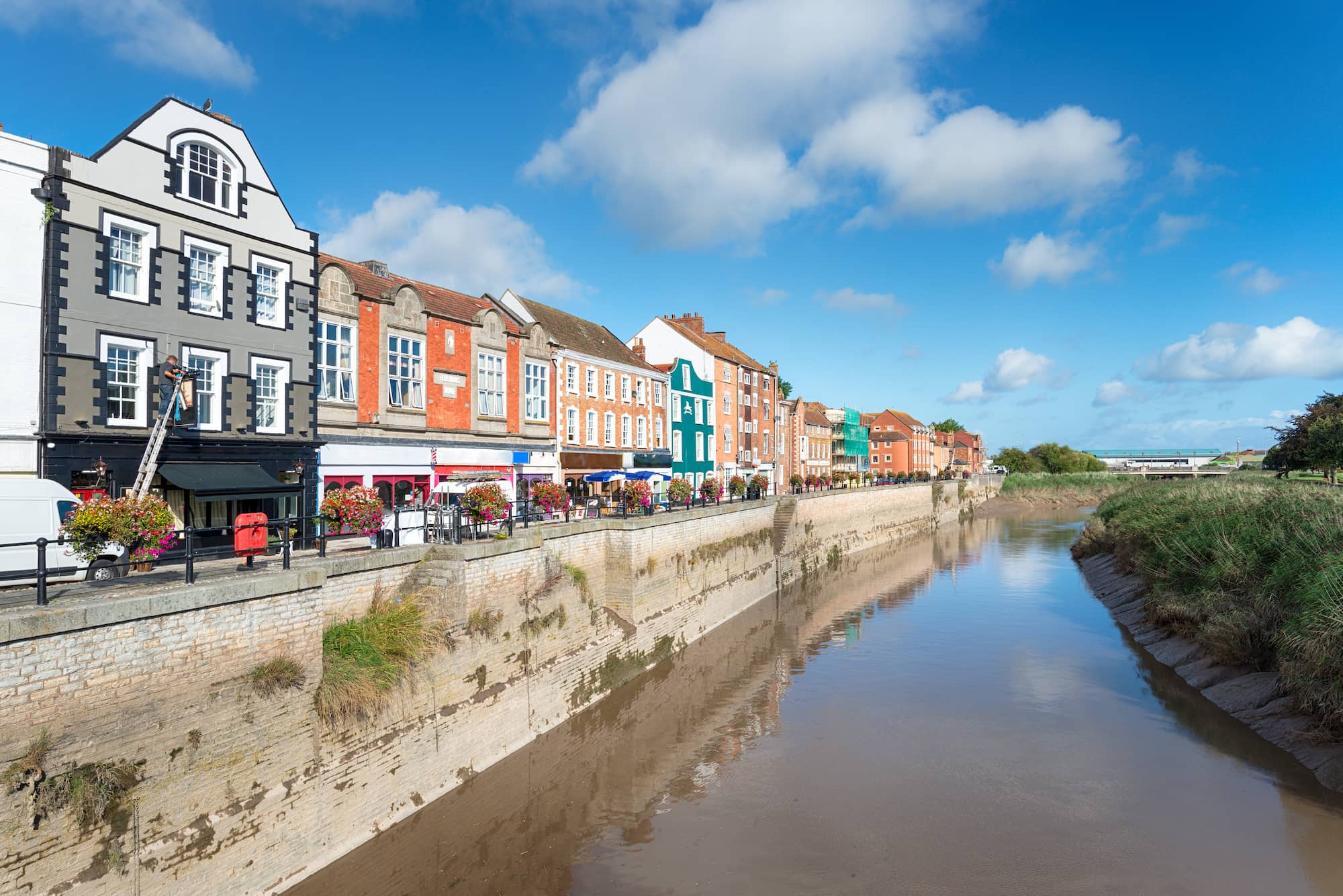 Colorful buildings line a riverside street under a blue sky with clouds. The calm river reflects the architecture, while green grass grows along the opposite bank.