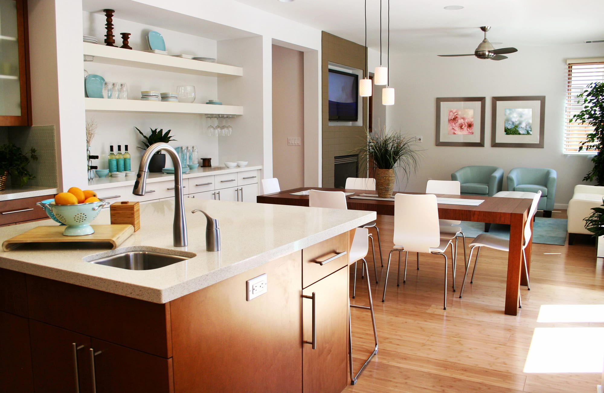 Modern open kitchen and dining area with wooden cabinets, a small sink, dining table with white chairs, and a cozy living space with pastel decor and large windows letting in natural light.